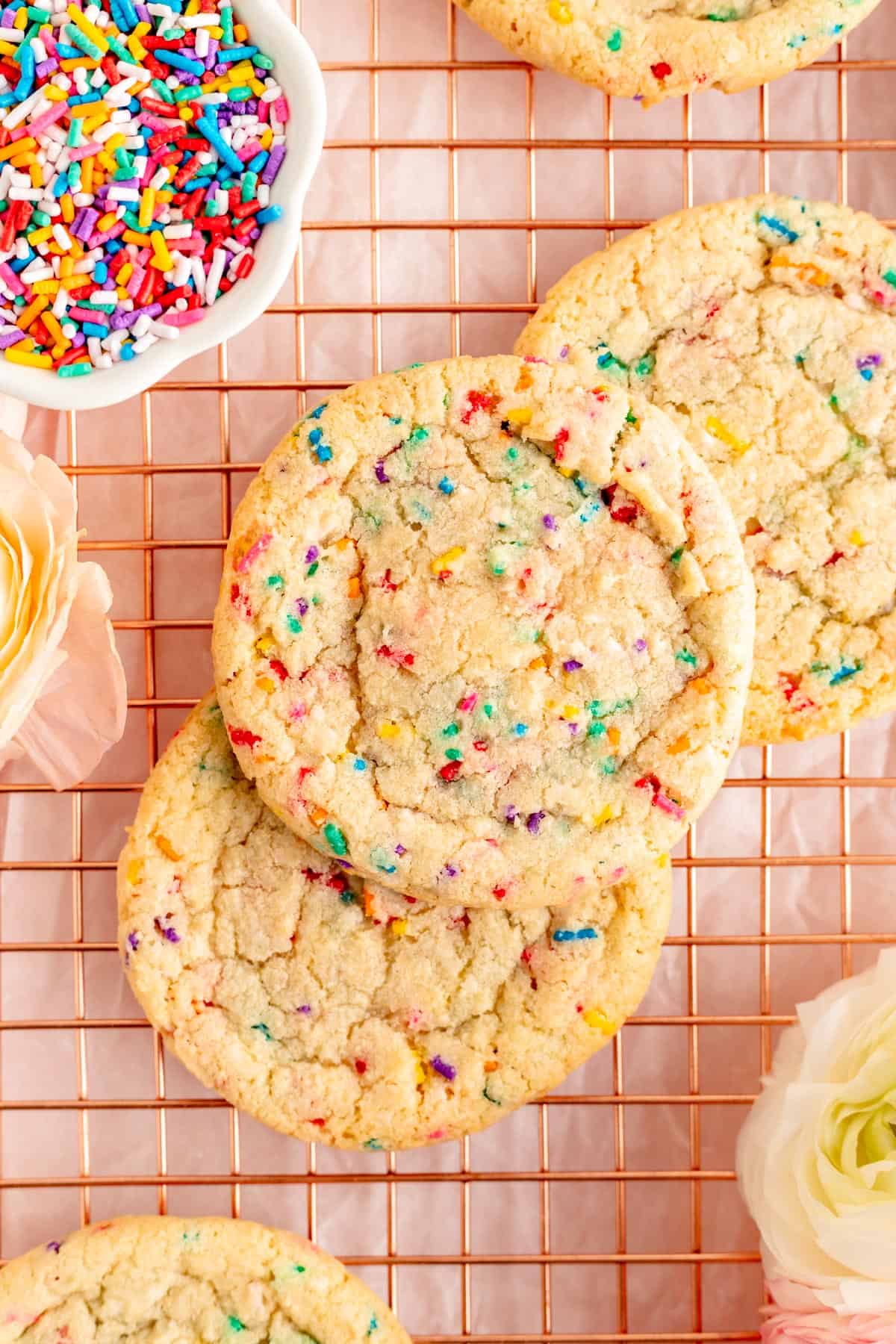 Close up of three funfetti cookies on copper wire rack with bowl of sprinkles and flowers.