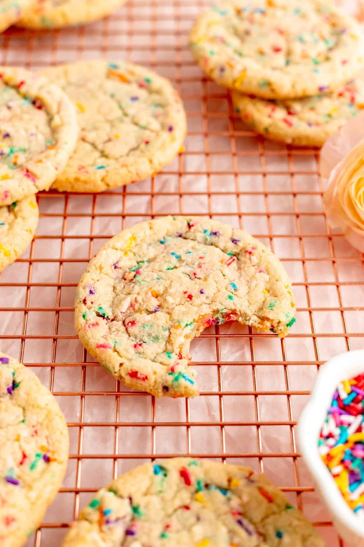 confetti sugar cookie with a bit out on copper wire rack surrounded by other sprinkle cookies.