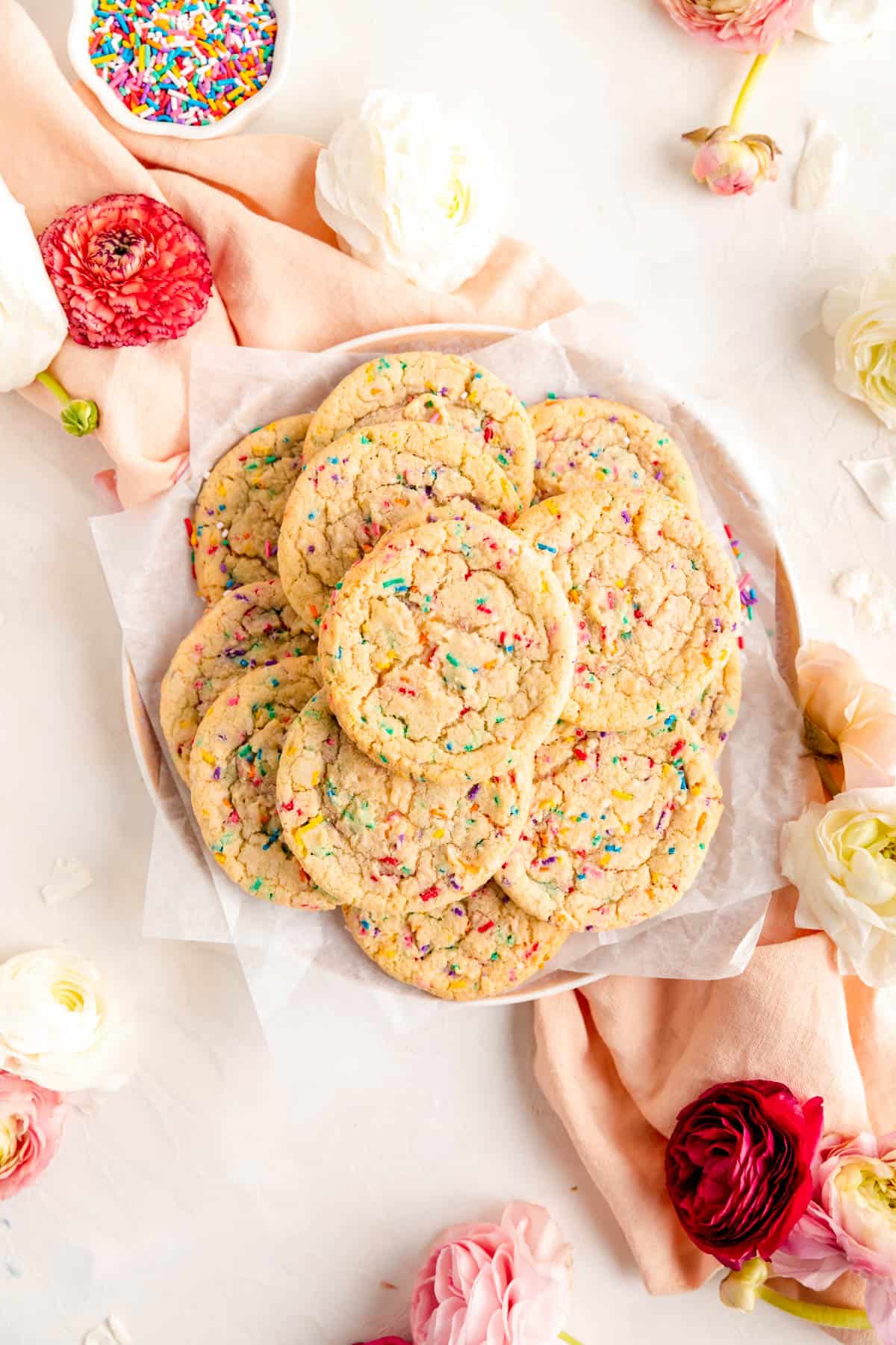 stack of funfetti cookies on a plate with pink linen and ranunculus on white background.