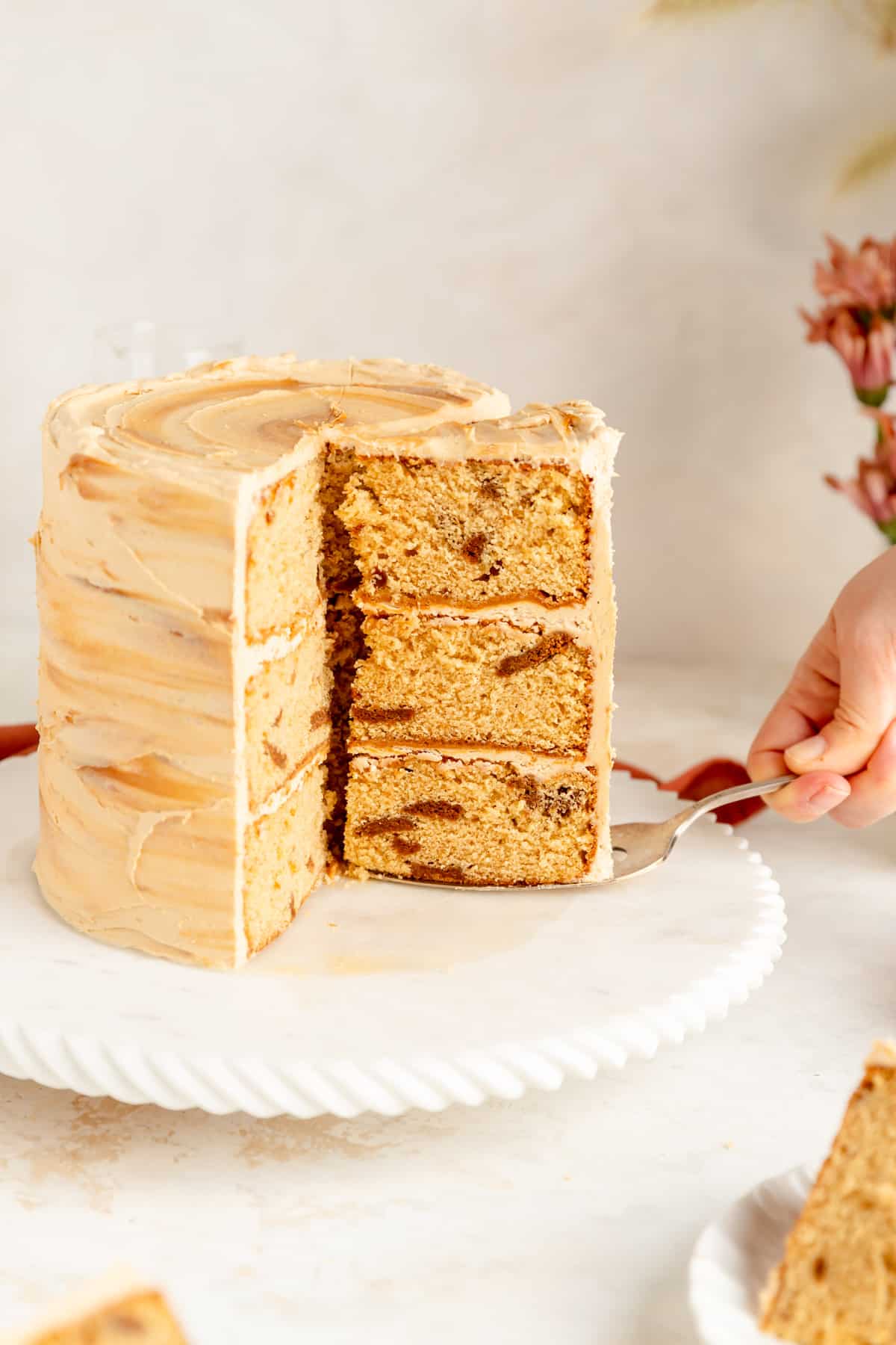 Hand using silver cake server to remove a slice of cookie butter cake from full cake.