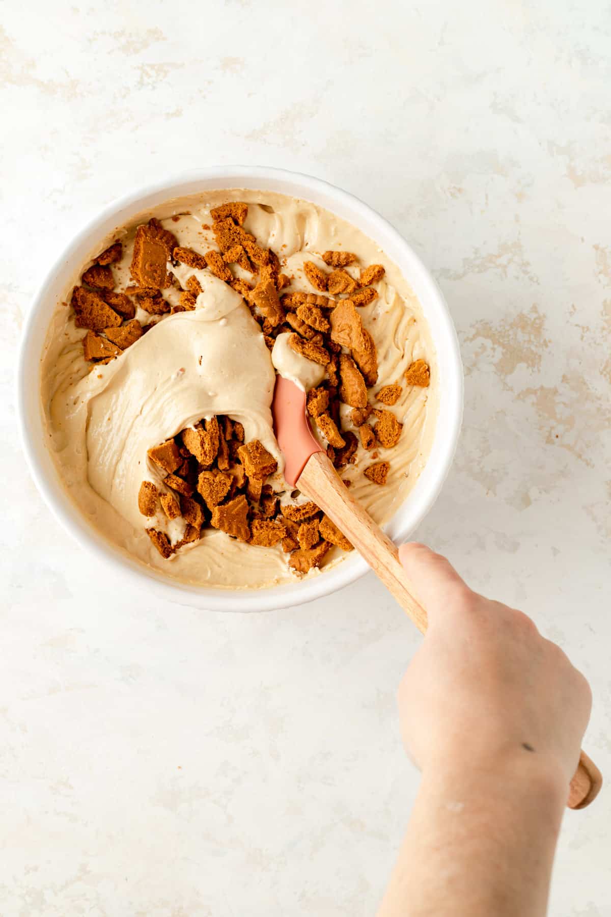 Hand holding wooden spatula folding crushed Biscoff cookies in to Biscoff cake batter.