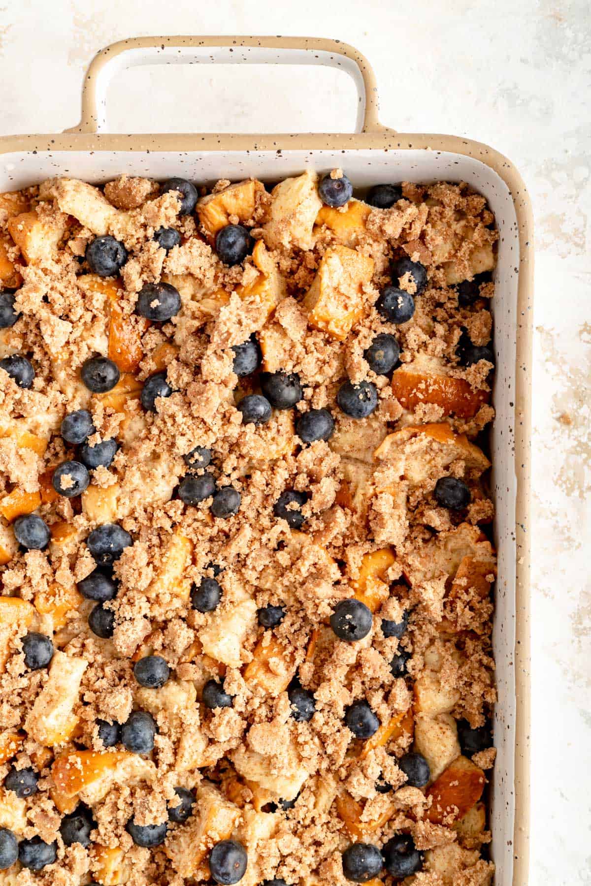 corner of a white and tan ceramic rectangular pan filled with bread chunks, blueberries, and streusel.