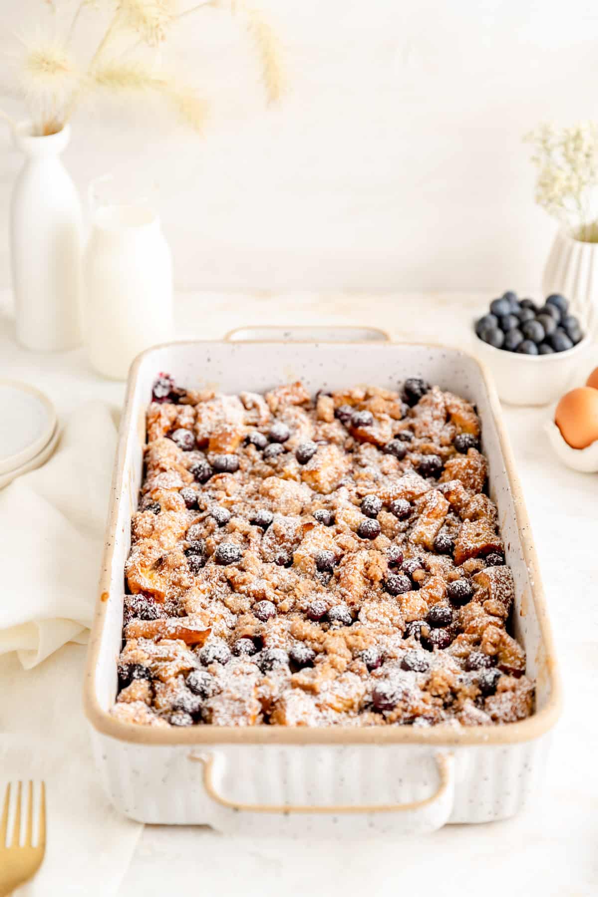 side angled shot of brioche french toast casserole in white ceramic pan with blueberries and vases.