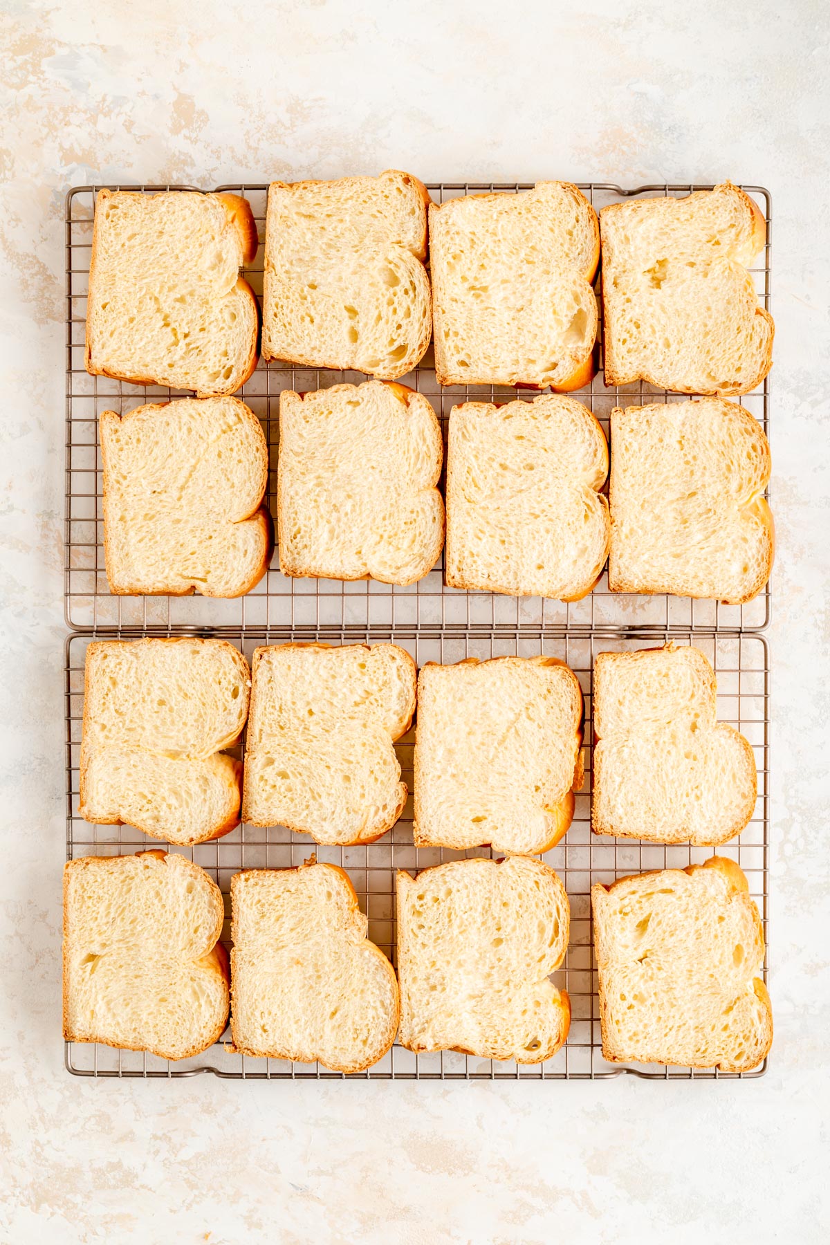 sixteen brioche bread slices spread out on cooling racks on white plaster background.