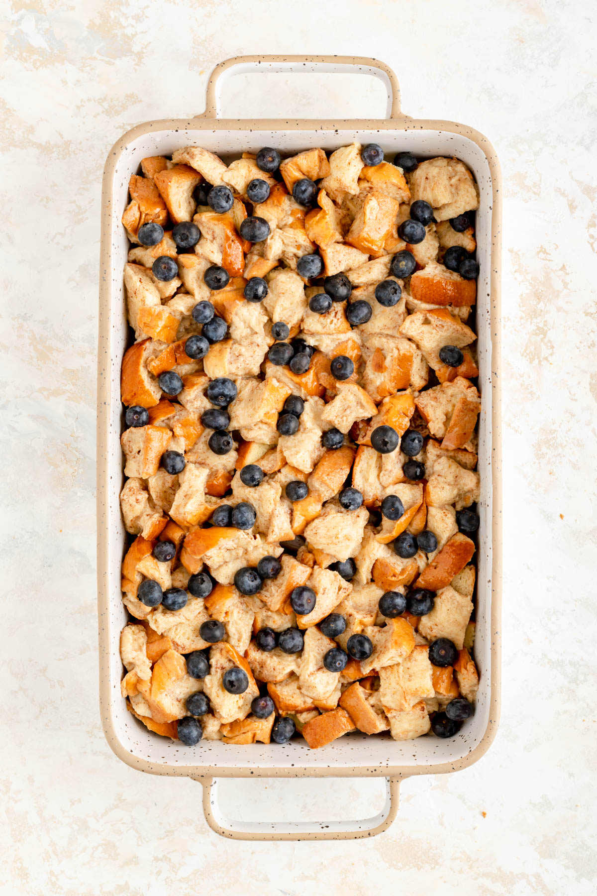 white and tan ceramic rectangular pan filled with bread pieces topped with fresh blueberries.