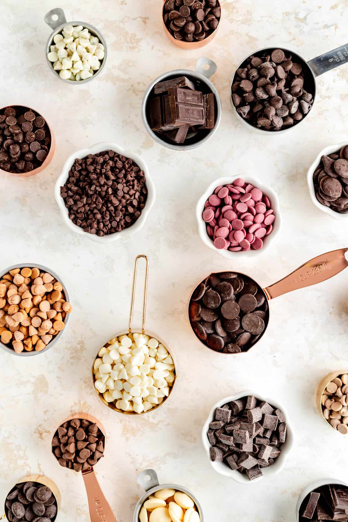 measuring cups filled with different colors and flavors of chocolate chips, pistoles, chunks, and bars.