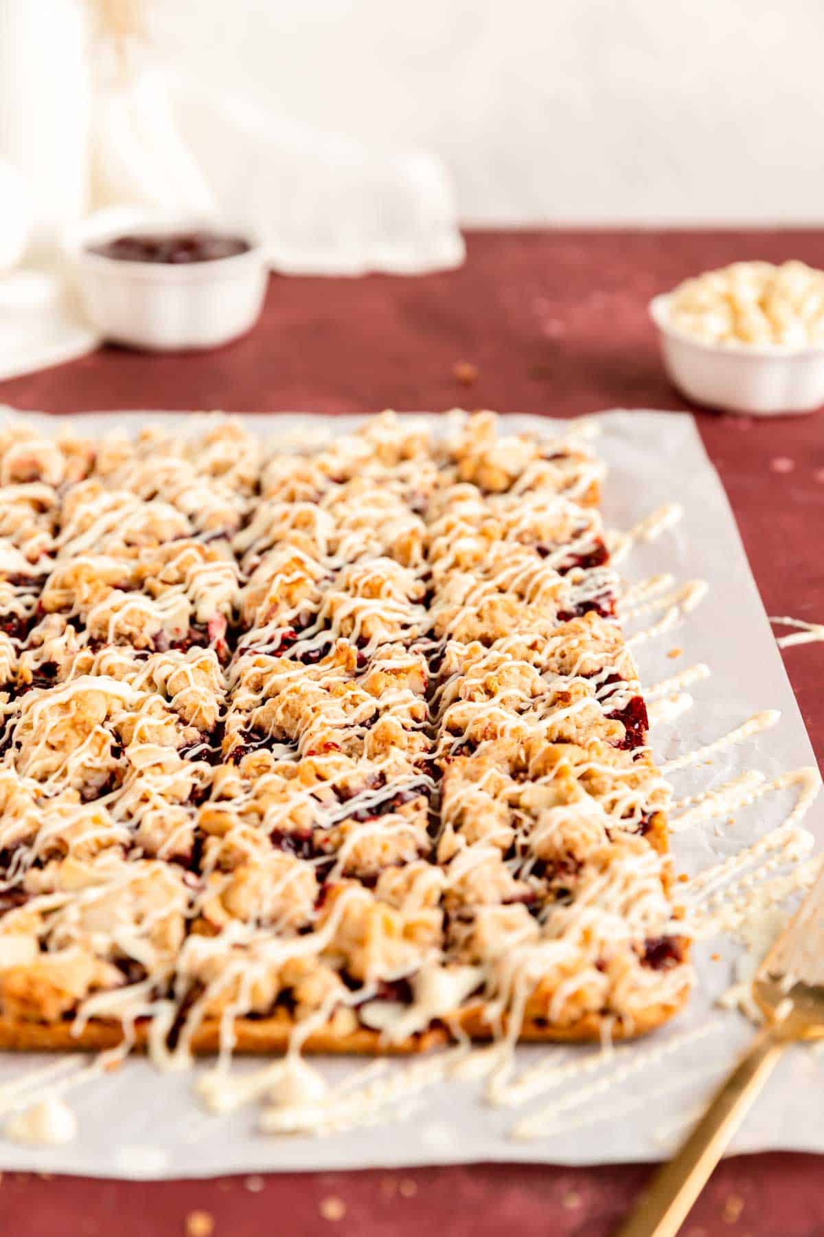 white chocolate drizzled raspberry jam bars on parchment with chocolate chips in background.