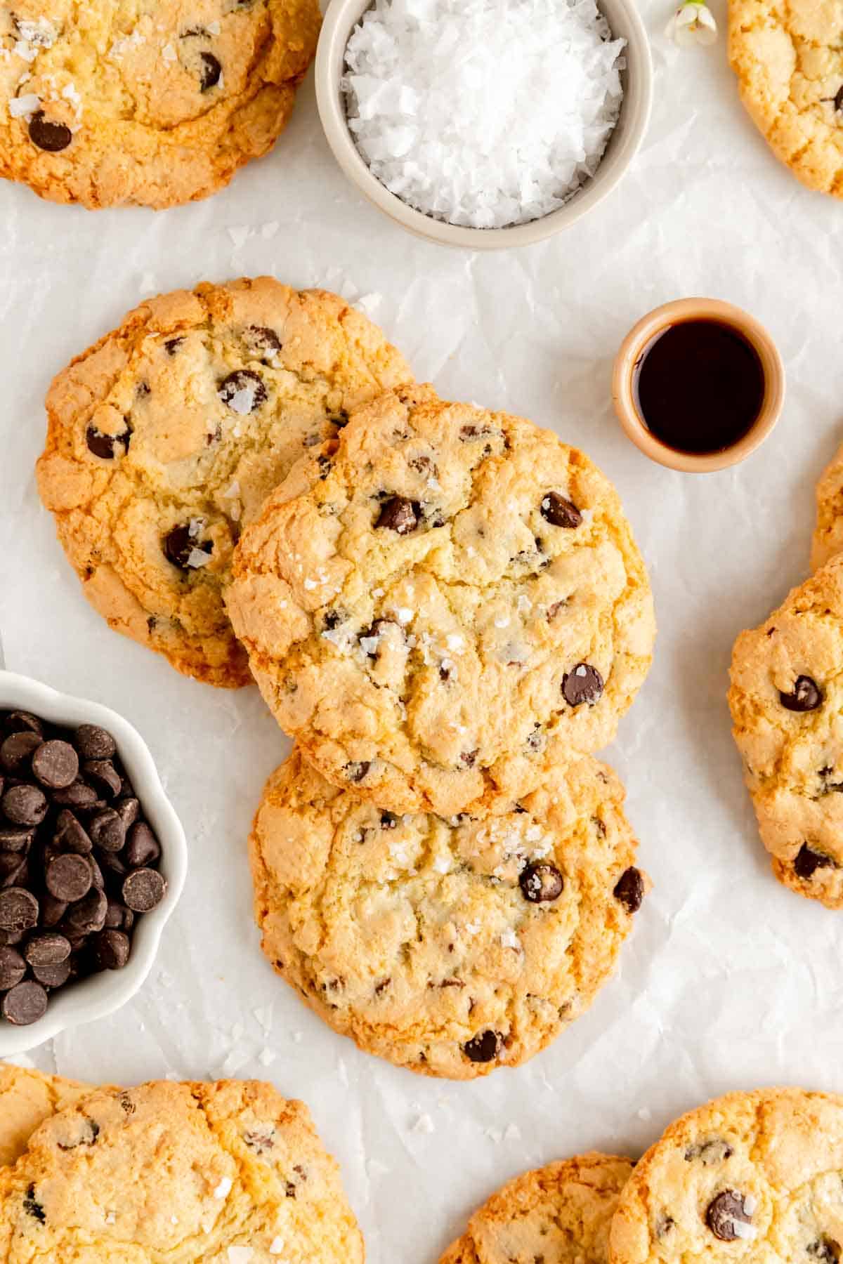 three chocolate chip cookies stacked on parchment paper surrounded by other cookies and ingredient bowls