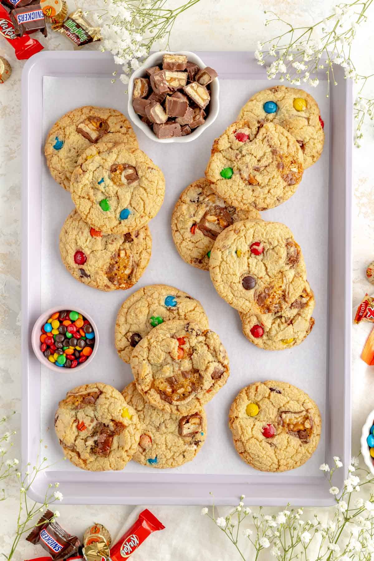 stacked candy bar cookies on a lavender baking tray with bowls of candy bits and pieces.