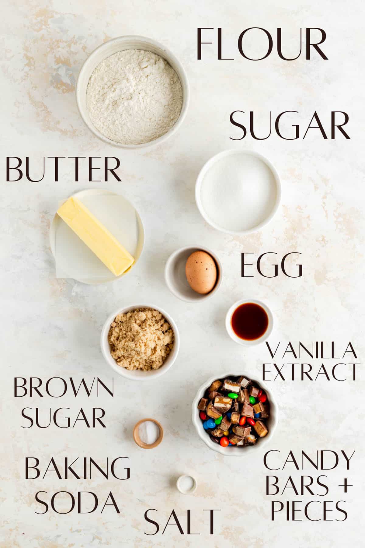 labeled ingredients for candy bar cookies in individual white bowls on white plaster background.